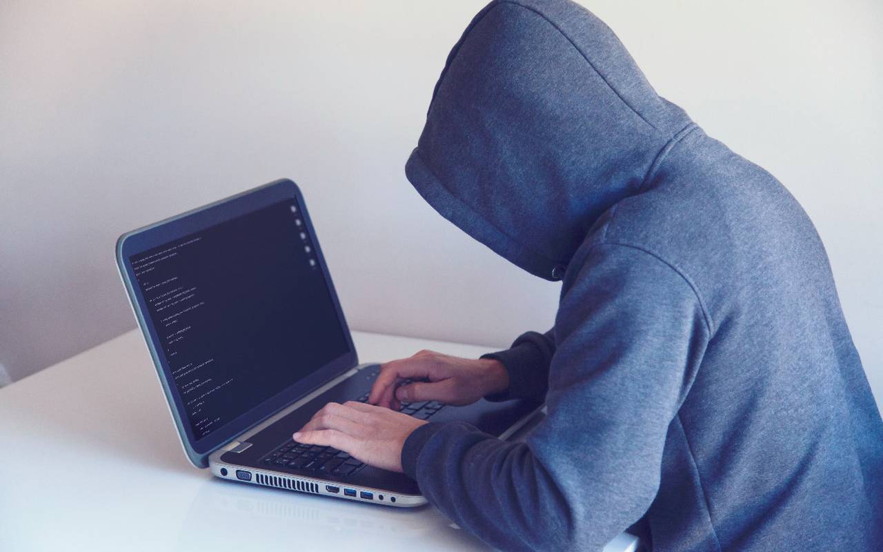 Eine Person sitzt vor einem Laptop. Sie hat einen grauen Kapuzen-Pulli an. Die Kapuze ist aufgezogen. Der Laptop steht auf einem weißen Tisch vor einer weißen Wand.