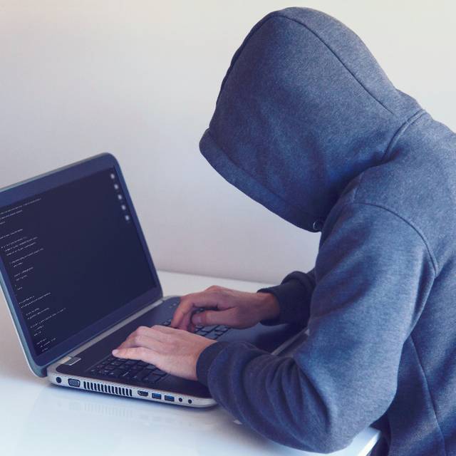 Eine Person sitzt vor einem Laptop. Sie hat einen grauen Kapuzen-Pulli an. Die Kapuze ist aufgezogen. Der Laptop steht auf einem weißen Tisch vor einer weißen Wand.