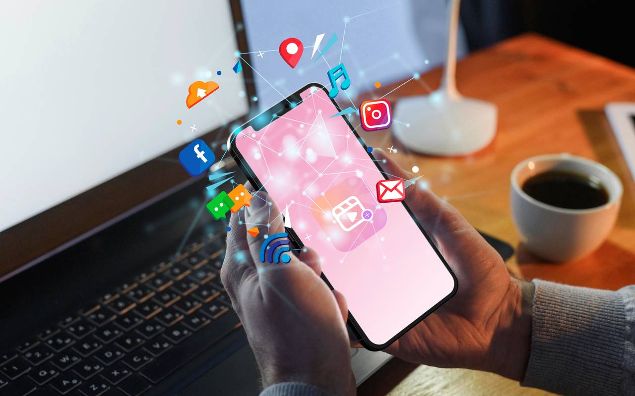 Jemand hält ein Handy in der Hand. Um das Handy herum sind Symbole von sozialen Medien und anderen Symbolen vom Handy