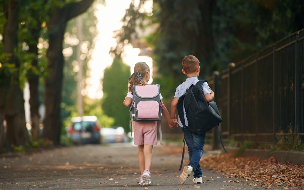 Ein Junge und ein Mädchen, mit Schulranzen auf dem Rücken halten sich an den Händen und laufen auf dem Gehweg