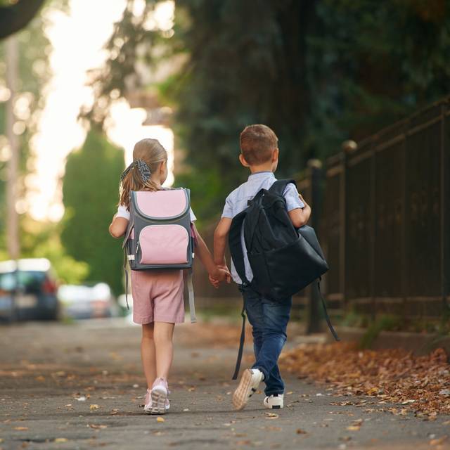 Ein Junge und ein Mädchen, mit Schulranzen auf dem Rücken halten sich an den Händen und laufen auf dem Gehweg