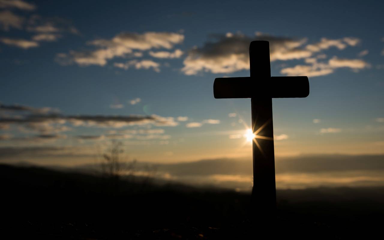 Ein Kreuz steht im Vordergrund. Im Hintergrund geht die Sonne unter.