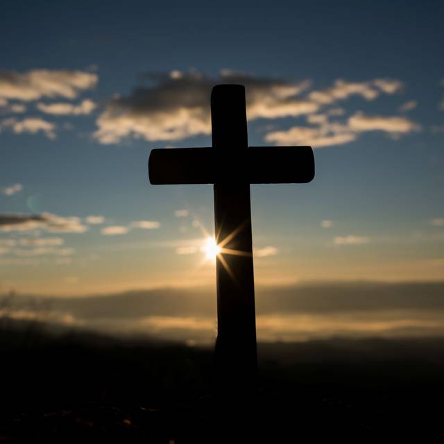 Ein Kreuz steht im Vordergrund. Im Hintergrund geht die Sonne unter.