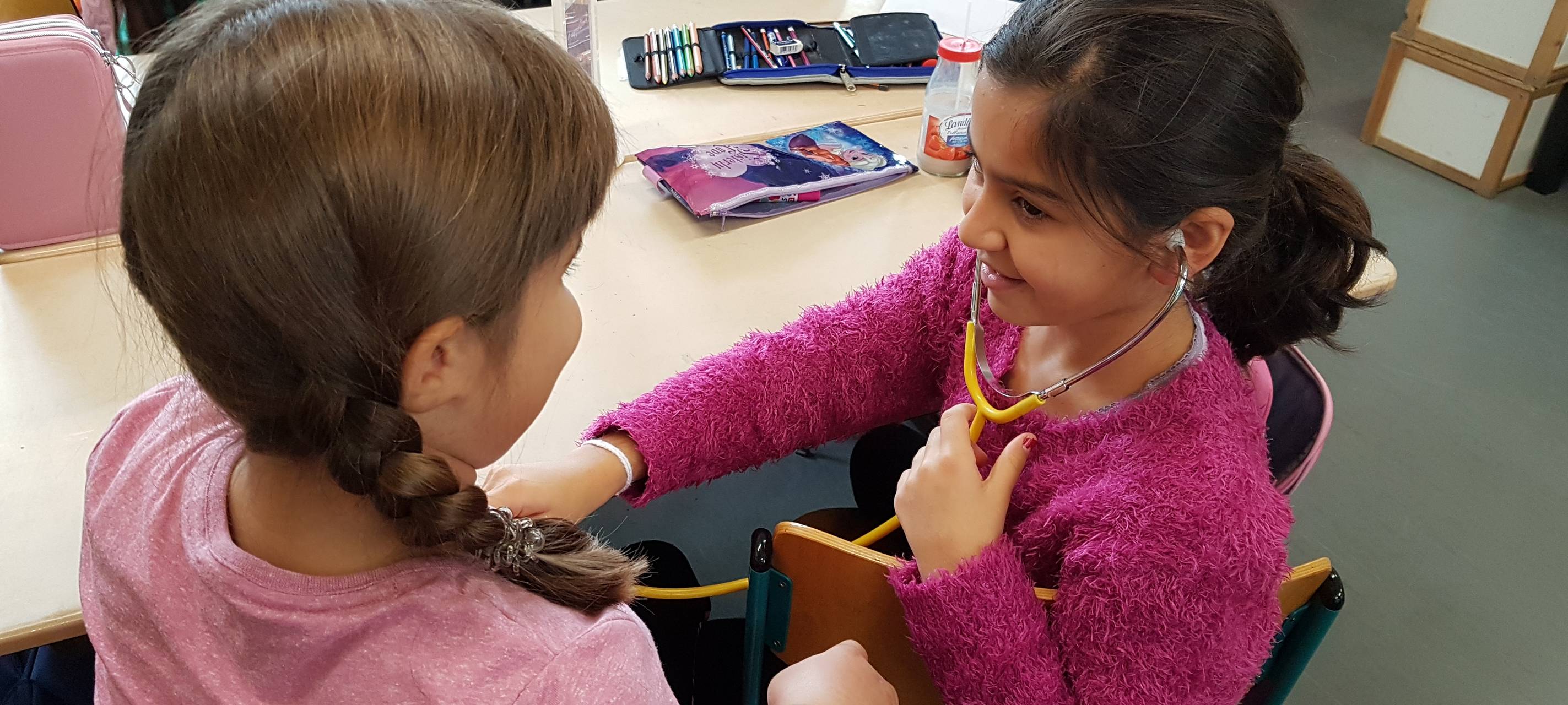 Zwei Mädchen nehmen am Unterricht "Klasse2000" teil und hören ihren Herzschlag an.