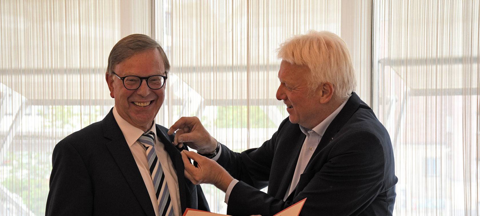 Zwei Männer im Anzug lächeln in die Kamera, einer steckt dem anderen eine Nadel an