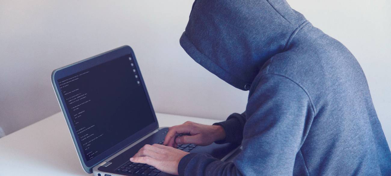 Eine Person sitzt vor einem Laptop. Sie hat einen grauen Kapuzen-Pulli an. Die Kapuze ist aufgezogen. Der Laptop steht auf einem weißen Tisch vor einer weißen Wand.