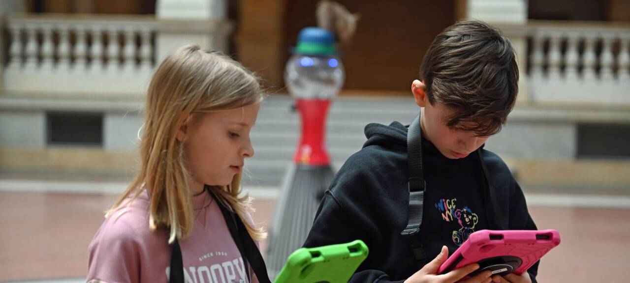 Linus (r) und Matilda stehen mit Tablets vor einem Roboter im Museum für Kommunikation Berlin.