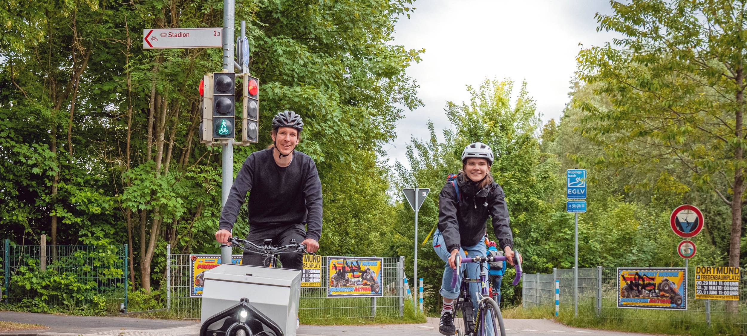 Symbolbild: Zwei Fahrradfahrer:innen fahren über die Straße und lächeln dabei in die Kamera.