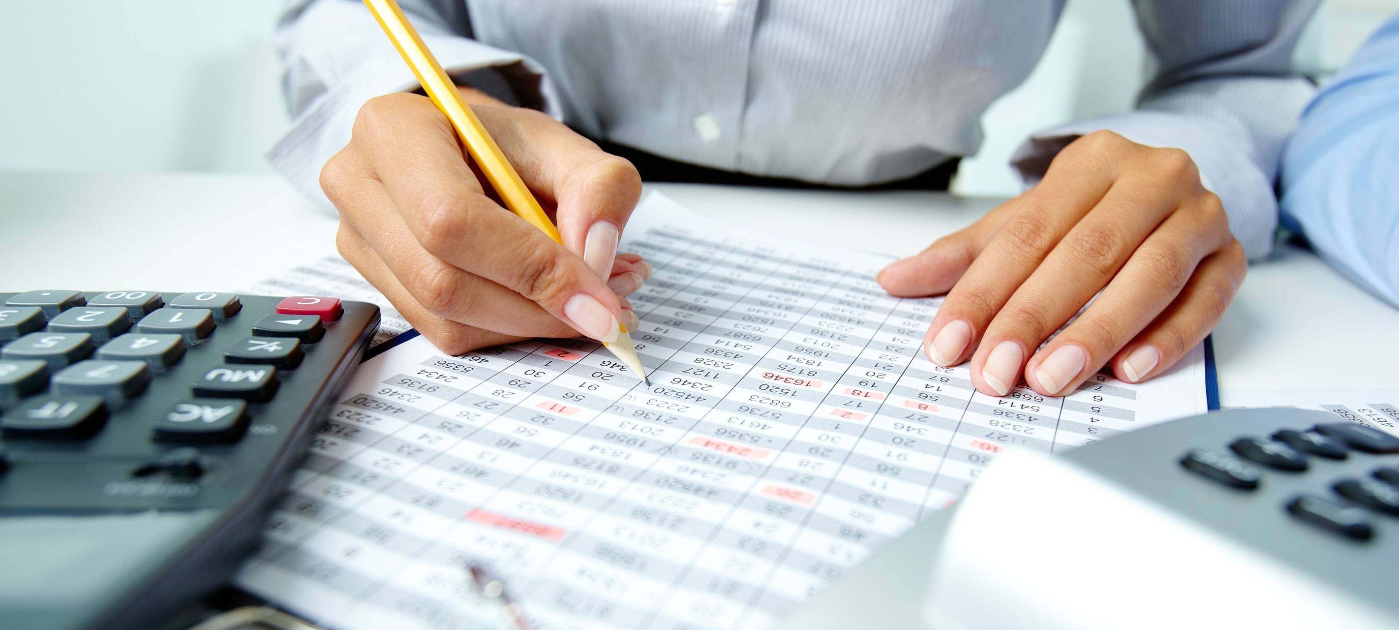 Unterlagen mit Zahlen und ein Taschenrechner auf dem Schreibtisch. Eine Frau geht die Unterlagen mit einem Stift durch