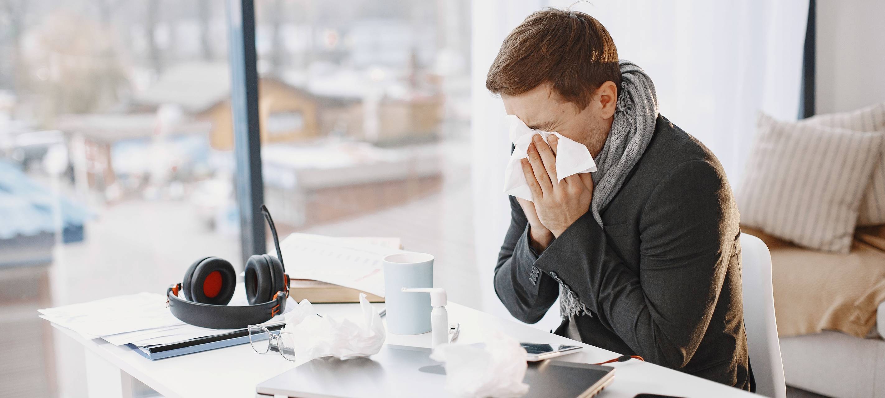 Ein Mann putzt sich am Schreibtisch die Nase. Auf dem Schreibtisch liegen Arbeitsmaterial und benutzte Taschentücher