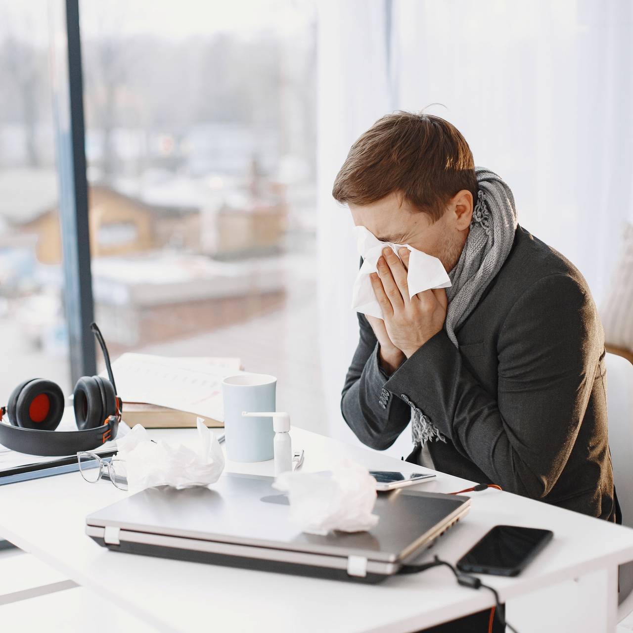 Ein Mann putzt sich am Schreibtisch die Nase. Auf dem Schreibtisch liegen Arbeitsmaterial und benutzte Taschentücher