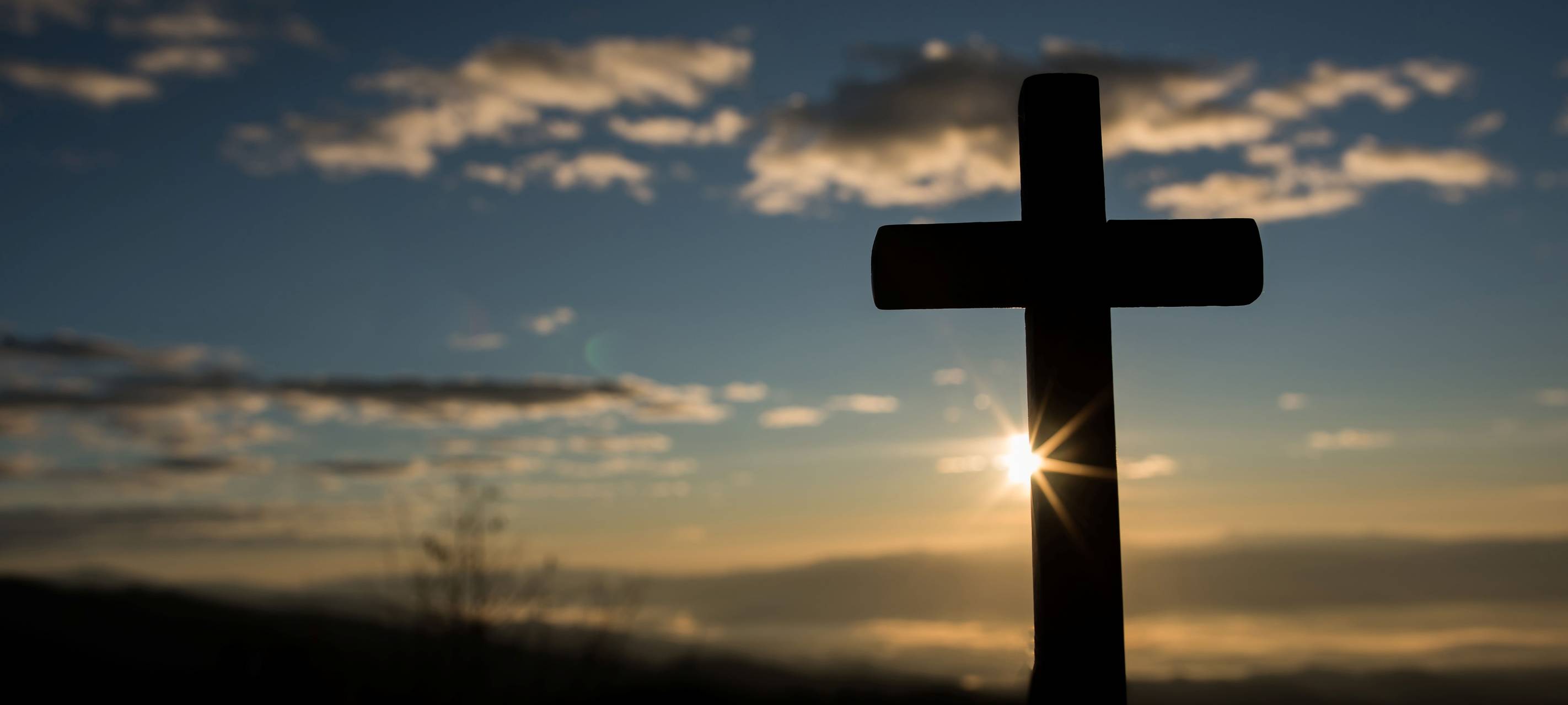 Ein Kreuz steht im Vordergrund. Im Hintergrund geht die Sonne unter.